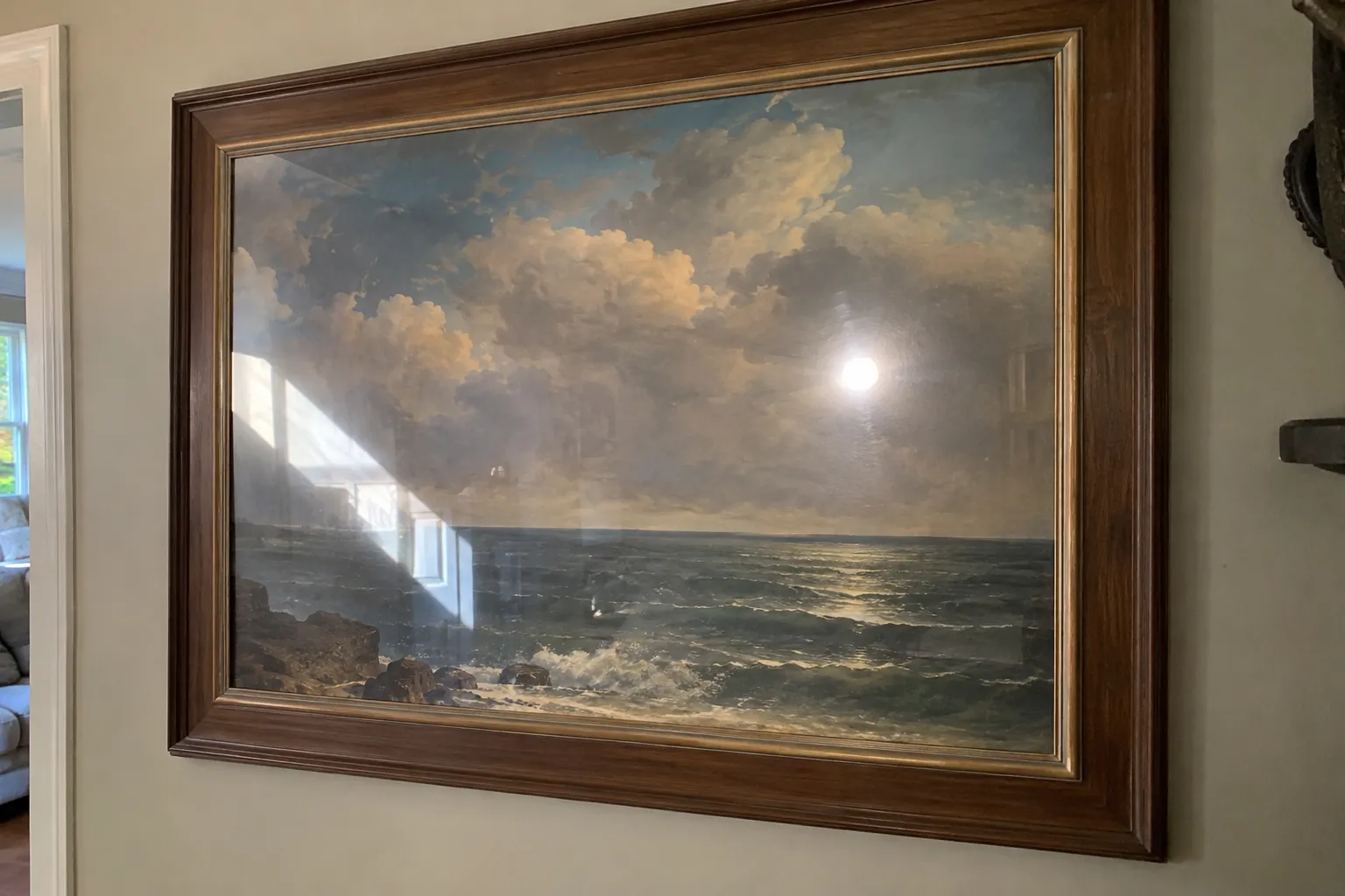 Framed seascape painting behind glass with a bright window reflection and a flash hotspot across it.