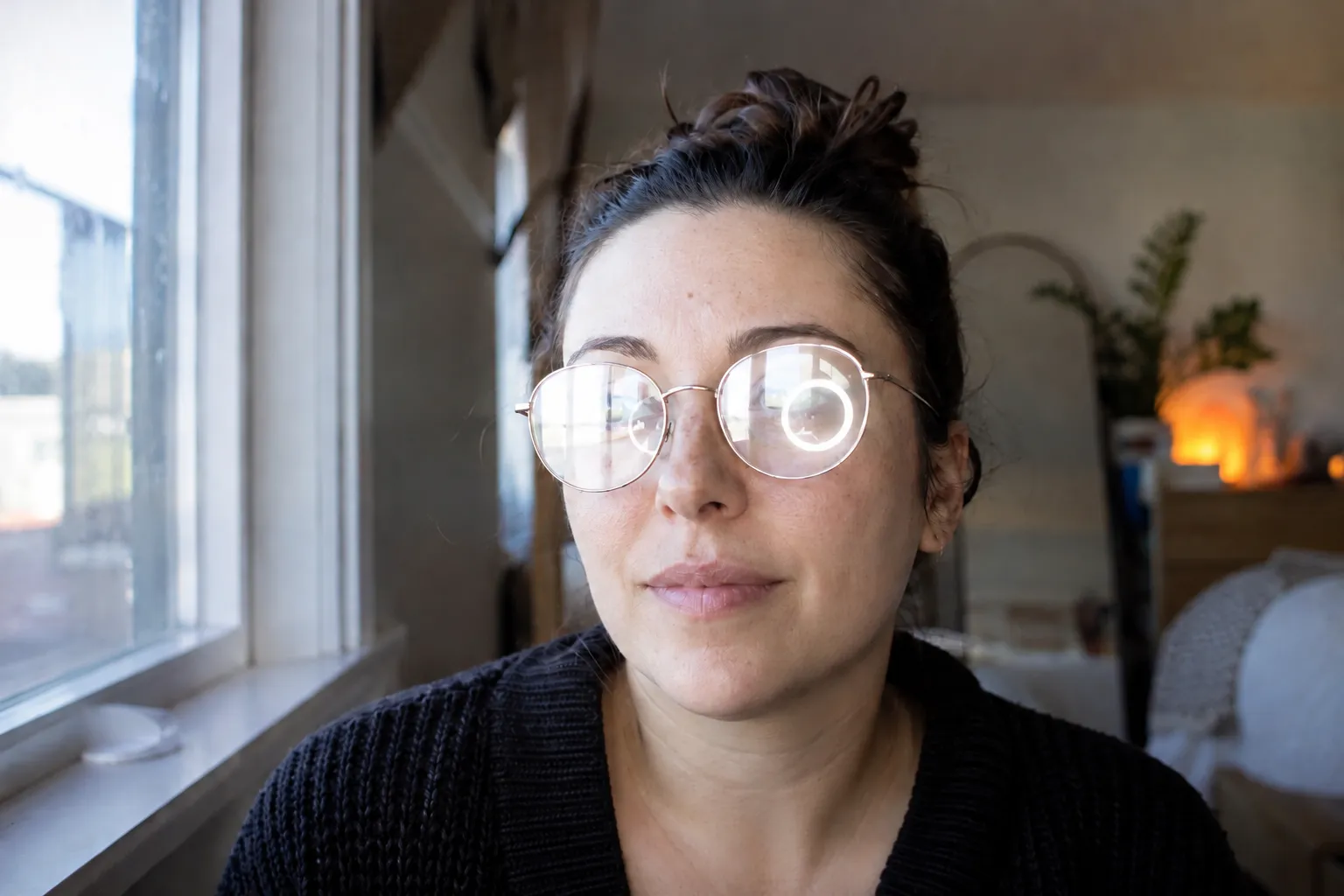Portrait of a woman whose eyeglass lenses reflect a bright window and a round ring light, hiding her eyes.